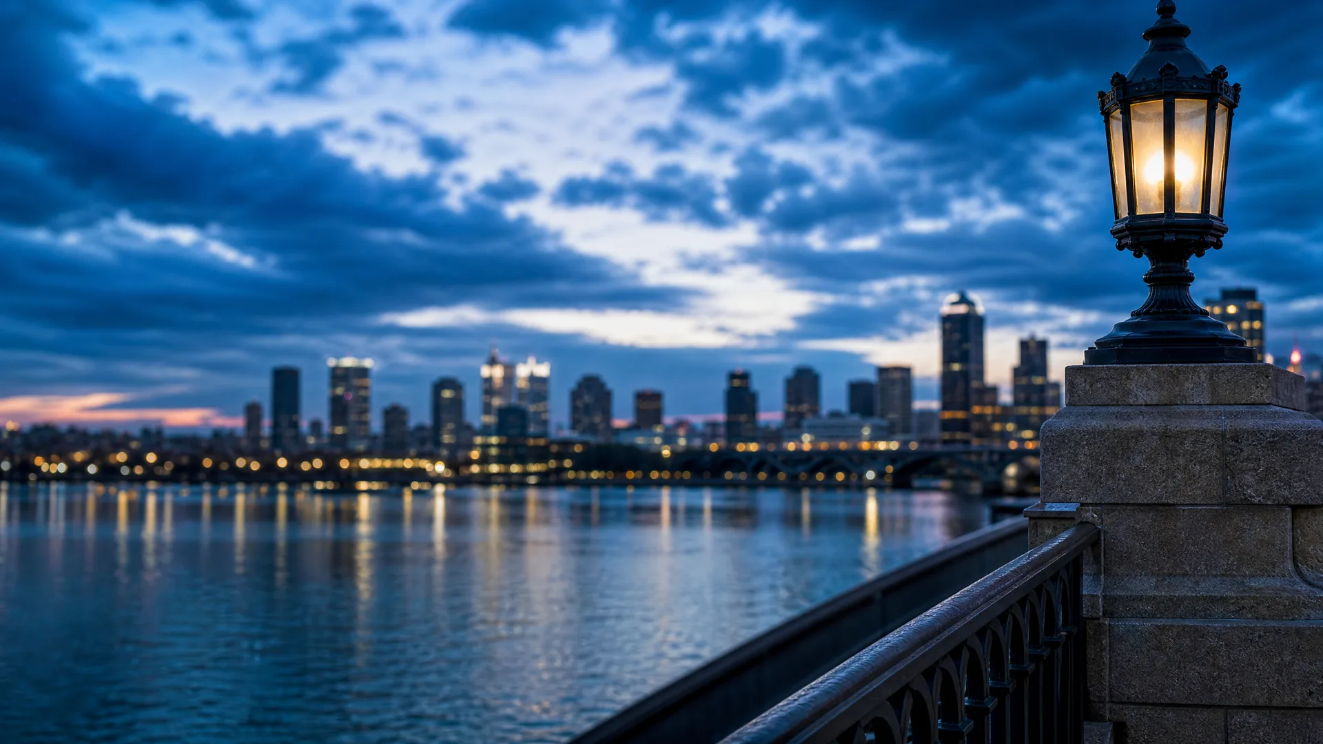 A lit ornate lamp on a stone pillar with an iron railing in the foreground, overlooking a wide river and a distant city skyline under heavy blue clouds at dusk.